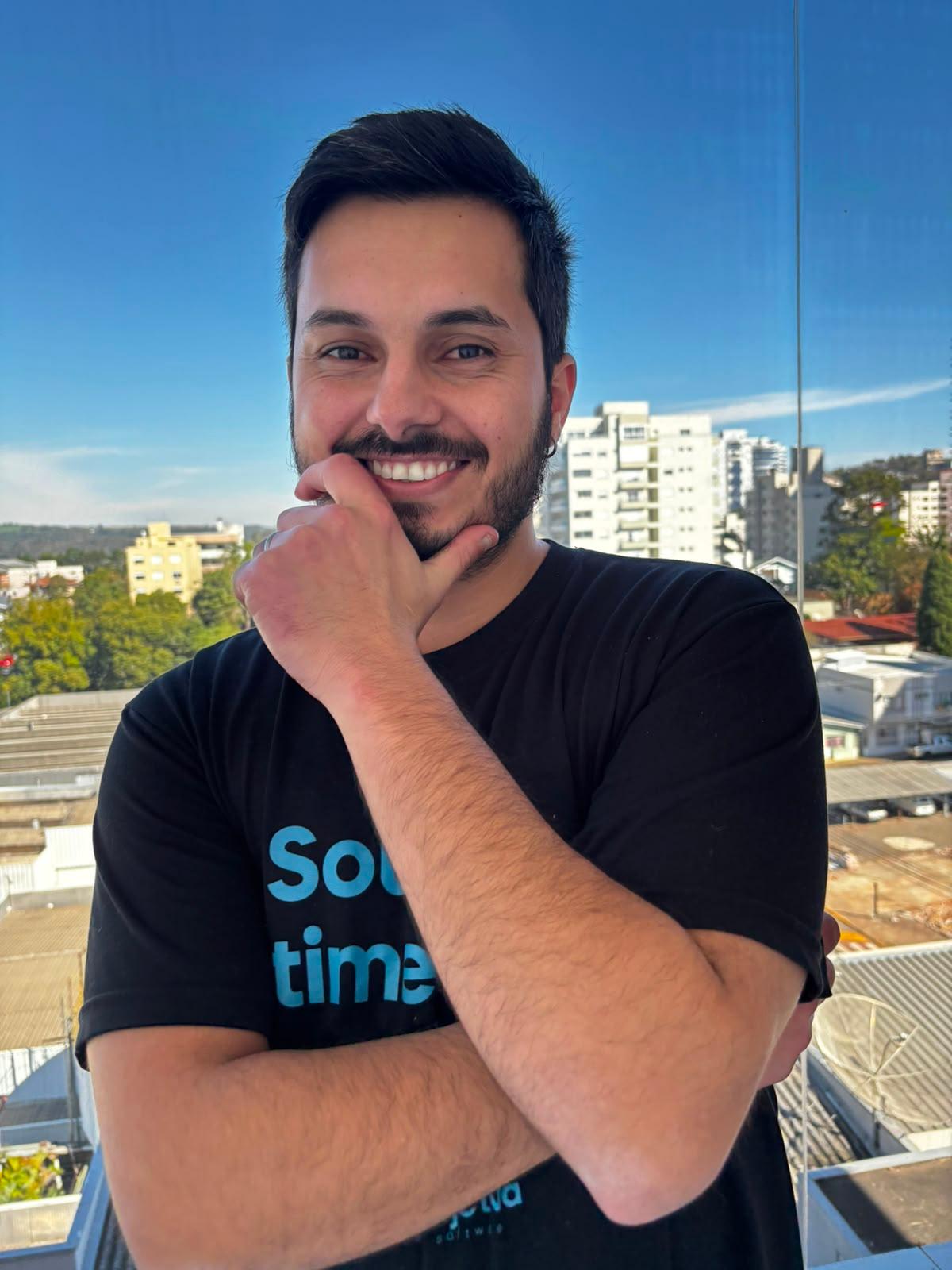 Matheus Maydana sorrindo, com a mão apoiada no queixo, em uma varanda com vista da cidade ao fundo.